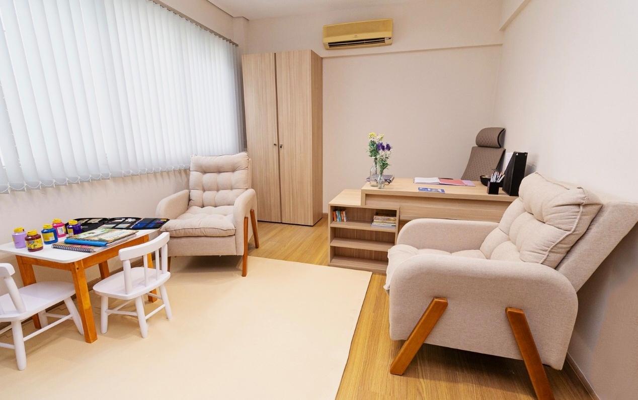 Sala de atendimento com duas poltronas, mesa de escritório em madeira clara e mesinha infantil com materiais de colorir