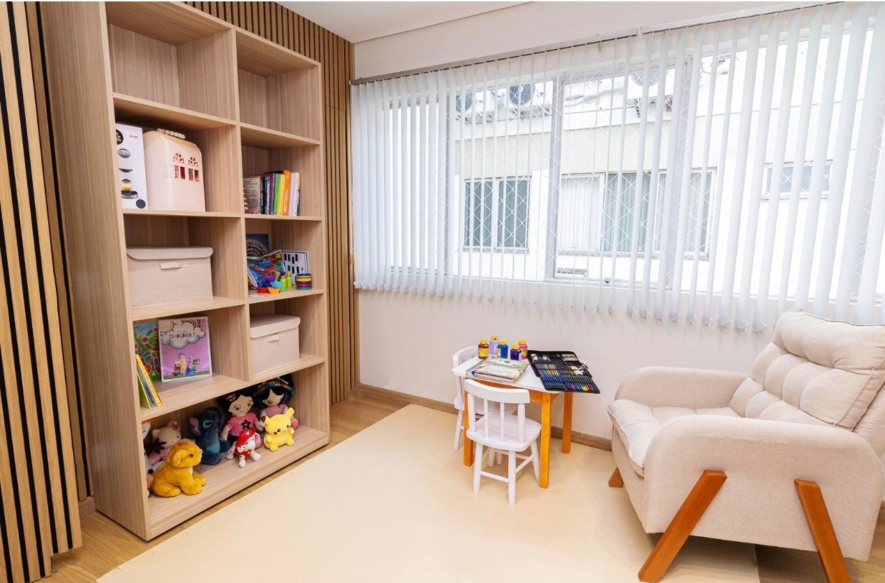 Sala de atendimento com estante de livros, jogos e brinquedos, mesa infantil com tintas e lápis e poltrona de atendimento