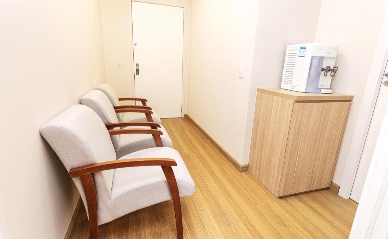 Espaço de espera com três poltronas estofadas, armário de madeira clara e bebedouro